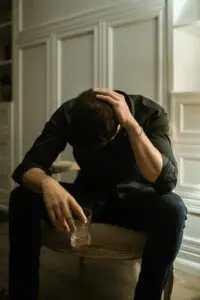 Man in distress seated indoors, head in hand, holding a drink, reflecting solitude.